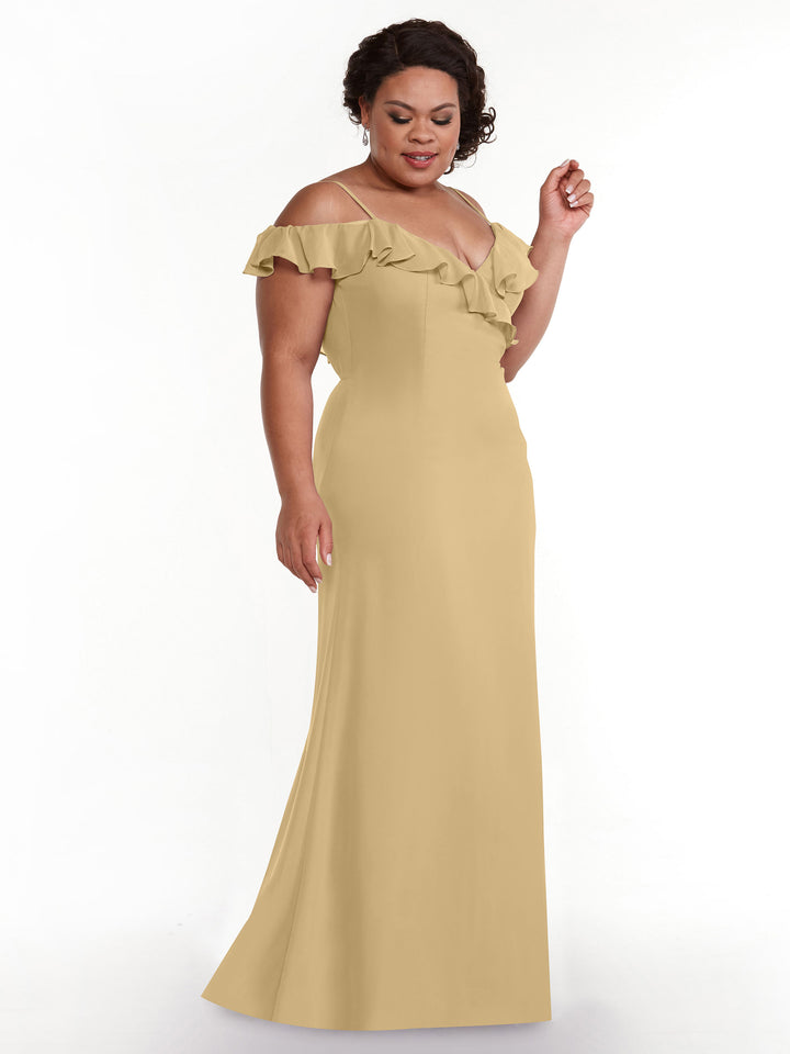 A woman in Avery Austin’s 80172 A-line, floor-length light gold gown with off-the-shoulder sleeves and a ruffled neckline stands against a white background, looking down with a gentle smile.