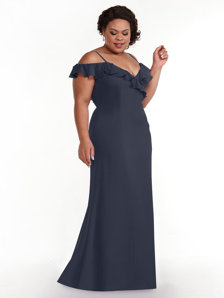 A woman in the Avery Austin 80172, a floor-length dark blue A-line dress with an off-the-shoulder ruffled neckline, stands against a plain white background, gazing down gently.