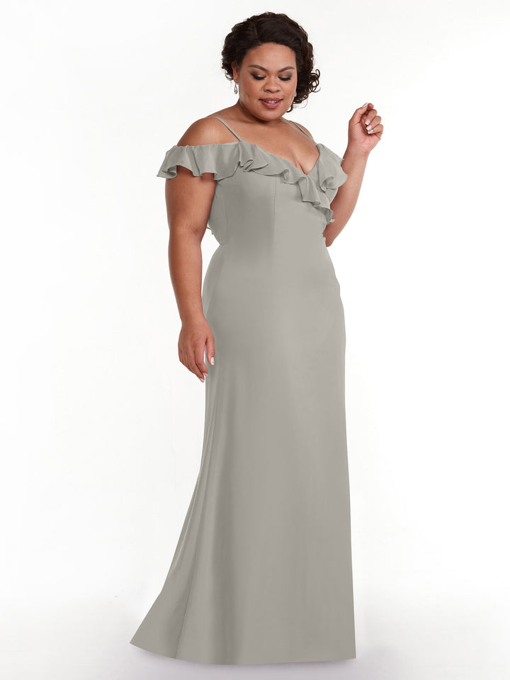 A woman in Avery Austin’s 80172, a light gray, floor-length A-line dress with ruffled neckline and off-the-shoulder sleeves, stands against a white background, gazing down gently.