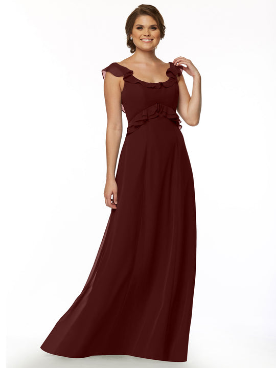 A woman wearing Avery Austin’s floor-length gown (Style 80173) with a ruffled neckline and ruffle accents at the shoulders and waist, standing and smiling against a plain white background.