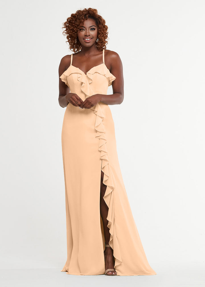 A woman with curly hair smiles against a white backdrop wearing Avery Austin’s 80174, a sleeveless light peach A-line dress with ruffle details on the sweetheart bodice and a diagonal ruffle trim accenting a high slit.