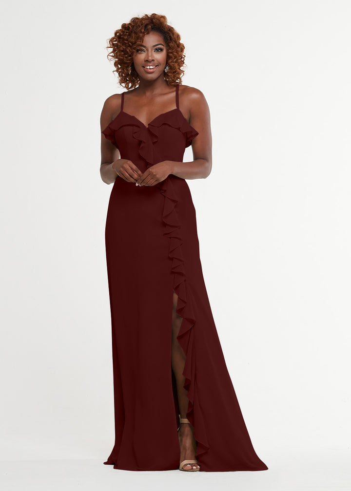 A woman with curly hair smiles against a white background, wearing Avery Austin’s sleeveless, floor-length ruffled dress (style 80174) featuring a sweetheart bodice and thigh-high slit.