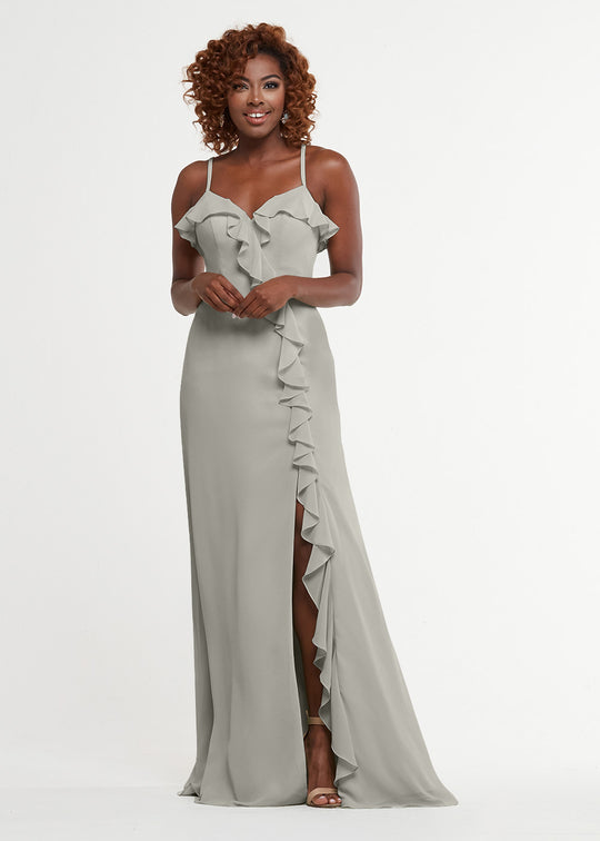 A woman with curly hair and heels smiles in front of a white background, wearing the Avery Austin 80174: a light gray, floor-length A-line dress featuring thin straps, ruffled neckline details, and a front slit.