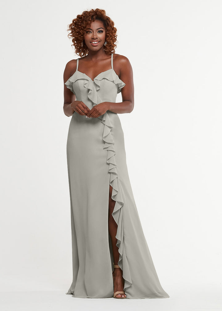 A woman with curly hair and heels smiles in front of a white background, wearing the Avery Austin 80174: a light gray, floor-length A-line dress featuring thin straps, ruffled neckline details, and a front slit.