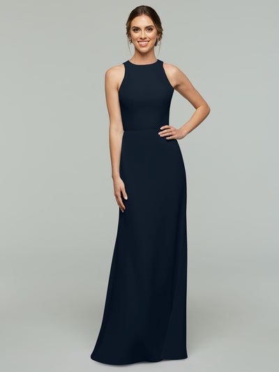 A woman, smiling with one hand on her hip and hair styled back, models Avery Austin’s sleeveless floor-length A-Line dress (style 80175) in dark navy blue against a plain gray background. The dress features a keyhole back.