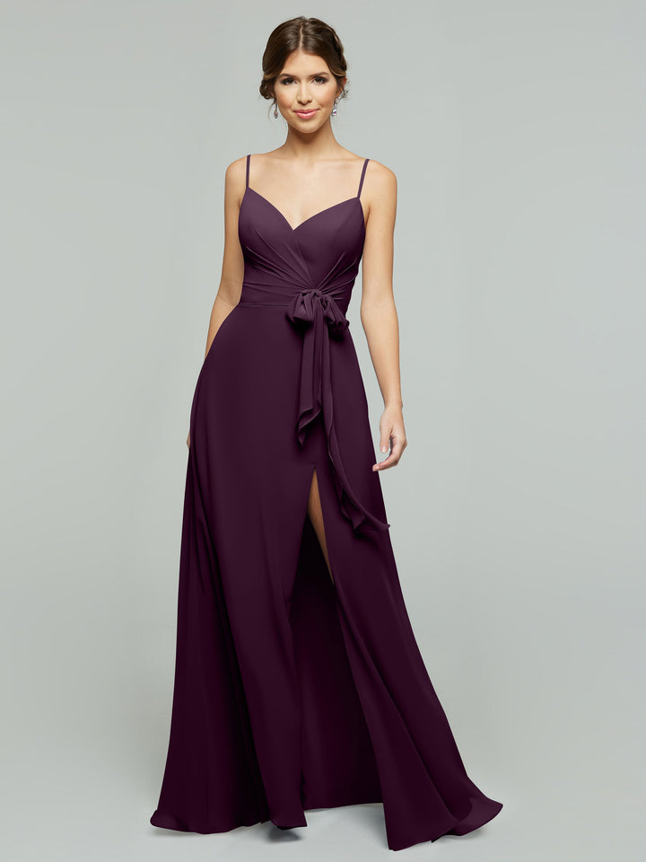 A woman wears the averyaustin 80182 floor-length Aubergine A-Line chiffon dress with thin straps, V-neck wrap bodice, waist tie, and high side slit, standing against a plain light gray background.