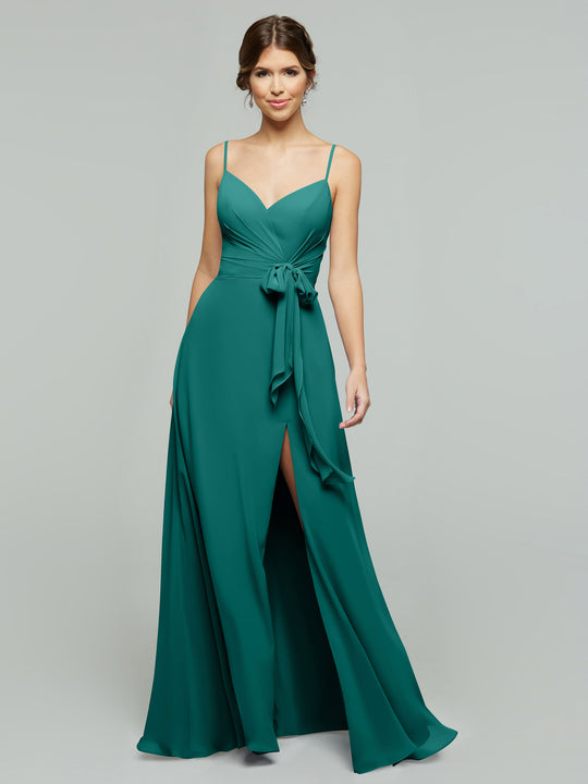 A woman wears averyaustin's 80182: a sleeveless, floor-length teal chiffon A-line dress with thin straps, v-neckline, side waist bow, and high skirt slit, standing before a plain light gray background.