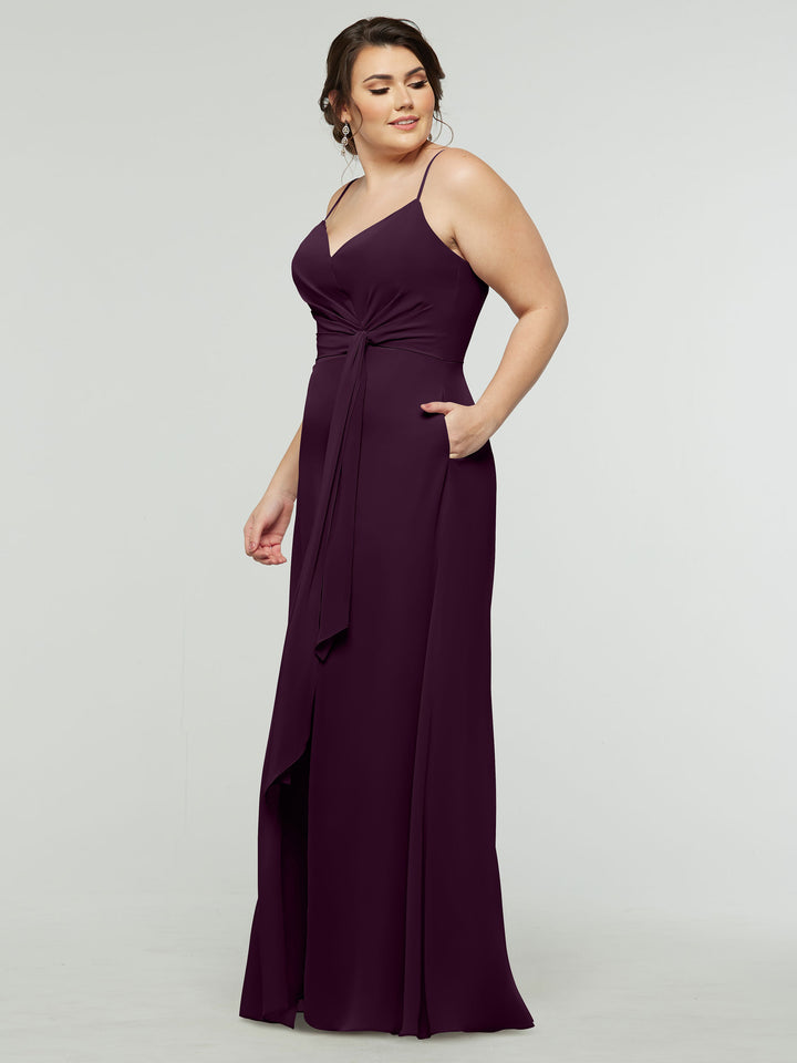 A woman in the averyaustin 80182 sleeveless aubergine A-line chiffon dress with V-neck, waist tie, side slit, and pockets stands against a plain light background, looking slightly to the side and smiling gently.