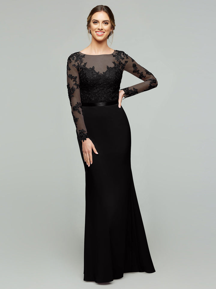 A woman models the averyaustin 80190 in Black, a long fitted gown with a lace bodice and illusion lace sleeves, standing against a plain light gray background and smiling with one hand on her hip.