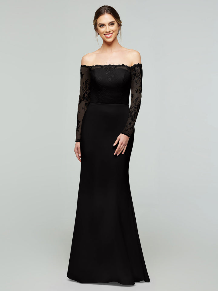A woman in a long, off-the-shoulder black gown (averyaustin 80191) with a lace bodice and sleeves stands smiling against a plain gray background. Her hair is styled back and she wears subtle earrings.
