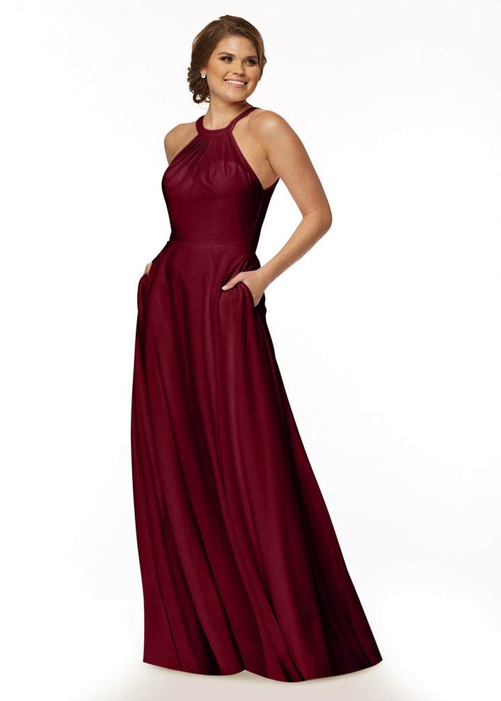 A woman stands smiling with her hands in the pockets of Avery Austin’s 83002, a sleeveless floor-length burgundy A-line dress with a high neckline, against a white background.