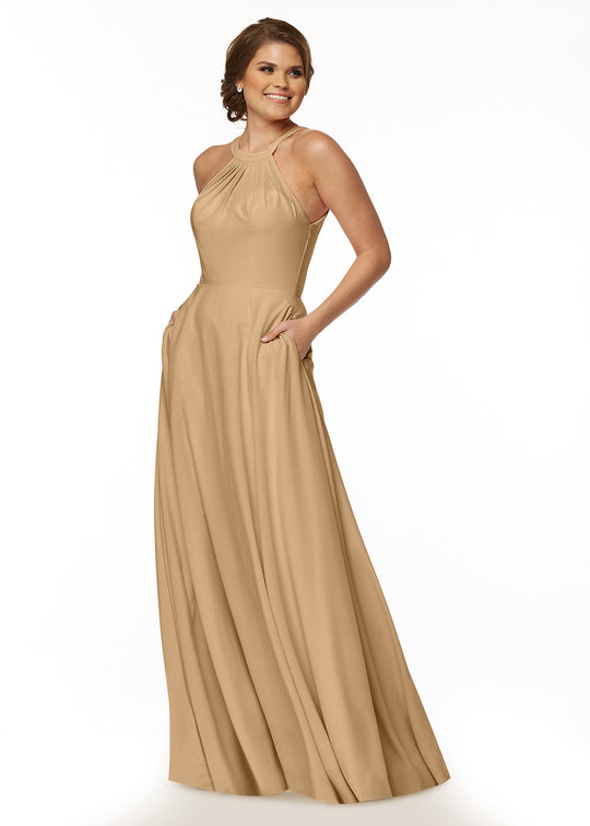 A woman smiles with her hands in the pockets of Avery Austin’s 83002—a long, sleeveless cappuccino A-line dress with a halter neckline—standing against a plain white background.
