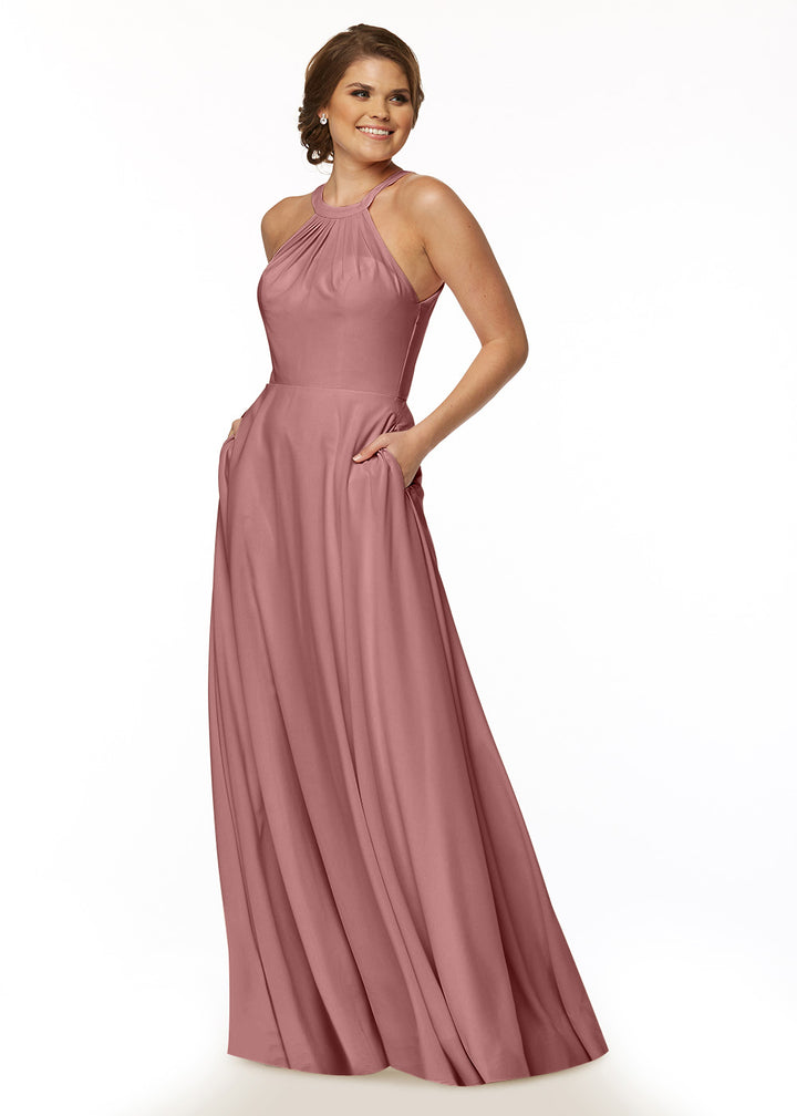 A woman smiles with hands in pockets, wearing Avery Austin’s floor-length A-line halter dress (style 83002) in dusty rose, featuring a sleeveless design and flowy skirt, set against a plain white background.
