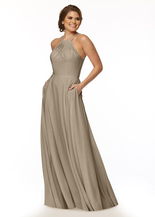 A woman stands smiling against a white background, wearing Avery Austin style 83002—a long taupe A-line dress featuring a halter neckline, flowy skirt with pockets.
