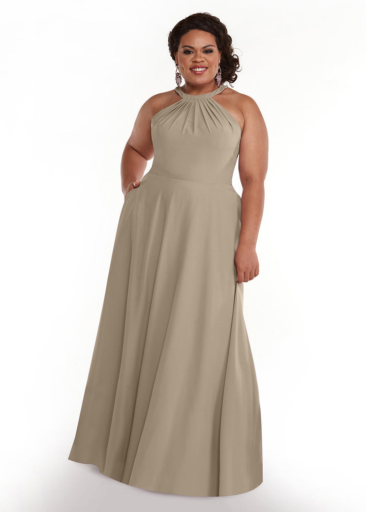 A woman stands and smiles against a plain white background, wearing Avery Austin style 83002—a sleeveless taupe halter neck gown with a gathered neckline and flowy floor-length skirt.