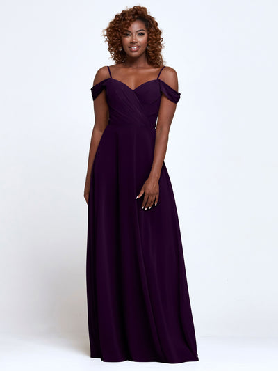 A woman poses against a plain backdrop in Avery Austin’s 83003—a deep purple, floor-length bridesmaid dress with thin straps and an off-the-shoulder fitted bodice. Her curly, shoulder-length hair adds to the elegant vibe as she smiles warmly.