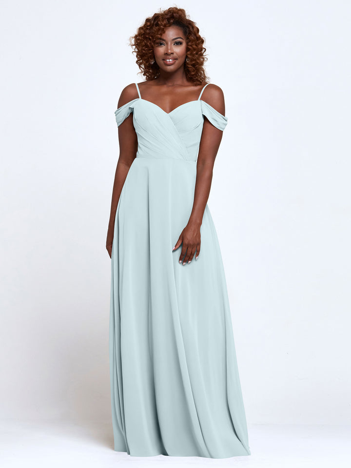 A woman with curly hair wears the Avery Austin 83003 Sky Blue off-the-shoulder bridesmaid dress featuring a gently pleated bodice and floor-length chiffon skirt, standing against a plain white background.