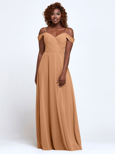 A woman with curly hair smiles at the camera while wearing Avery Austin’s floor-length bridesmaid dress (Style 83003) in tan chiffon, featuring an off-the-shoulder neckline and draped fabric, standing against a plain white background.