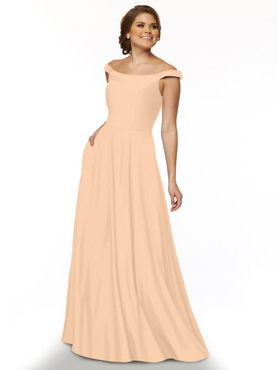 A woman smiles with one hand in her pocket, modeling the Avery Austin 83005 peach off-the-shoulder dress with a straight-across neckline against a plain white background.