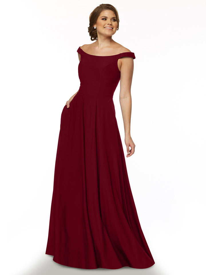 A woman smiles in front of a plain white background wearing the Avery Austin 83005 dress, a floor-length burgundy off-the-shoulder gown with a fitted bodice and flowing skirt, her hand stylishly tucked in her pocket.