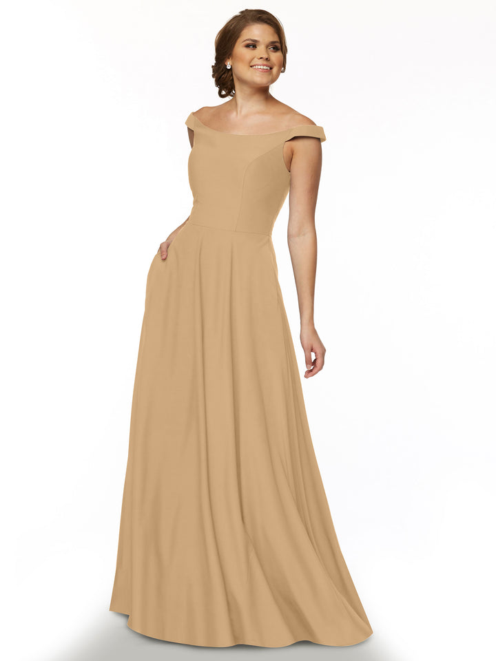 A woman smiles with one hand in her pocket, modeling Avery Austin’s 83005 dress in cappuccino. The off-the-shoulder, straight neckline elegantly showcases her shoulders against a plain white background.