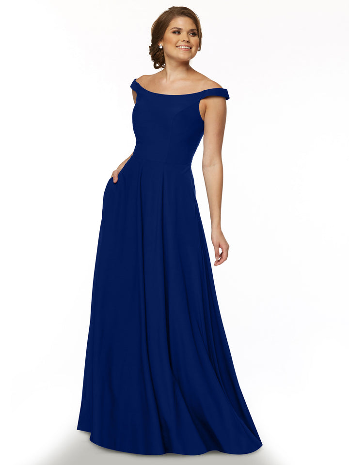 A woman models Avery Austin’s 83005: a straight across, off-the-shoulder, floor-length royal blue gown with pockets, standing against a white background and smiling slightly.