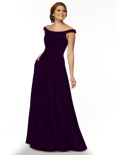 A woman smiles with one hand in her pocket, modeling Avery Austin style 83005—a deep purple, off-the-shoulder dress with a straight-across neckline—against a plain white background, ready to show off shoulders effortlessly.