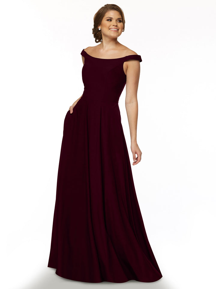 A woman smiles and poses gracefully in the Avery Austin 83005, a maroon floor-length, off-the-shoulder dress with a straight-across neckline and flowing skirt, set against a plain white background.