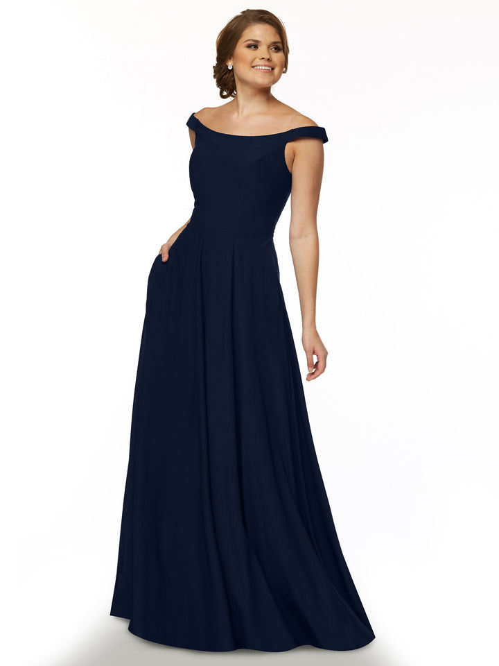 A woman smiles in the Avery Austin TH-83005 gown in Midnight, standing with one hand in her pocket to highlight its straight across, off-the-shoulder design against a plain white background.
