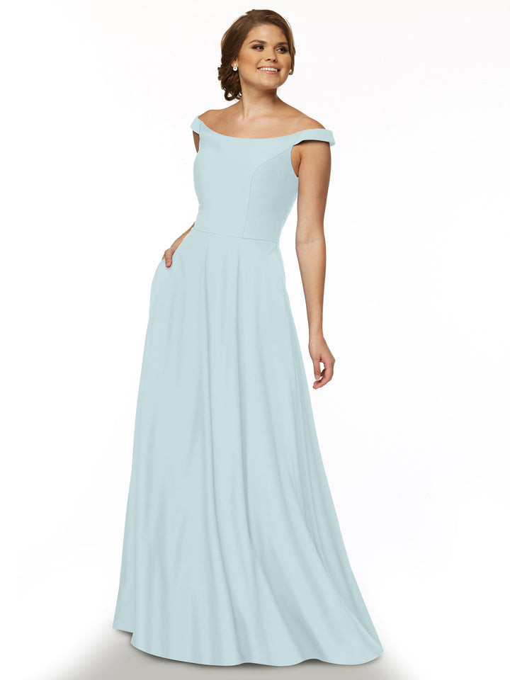 A woman in a sky blue, off-the-shoulder Avery Austin dress (style 83005) stands against a white background, smiling with one hand in her pocket.