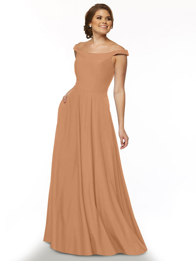 A woman models Avery Austin’s 83005, a floor-length off-the-shoulder dress with a straight neckline, standing against a white background and smiling slightly with one hand in her pocket.