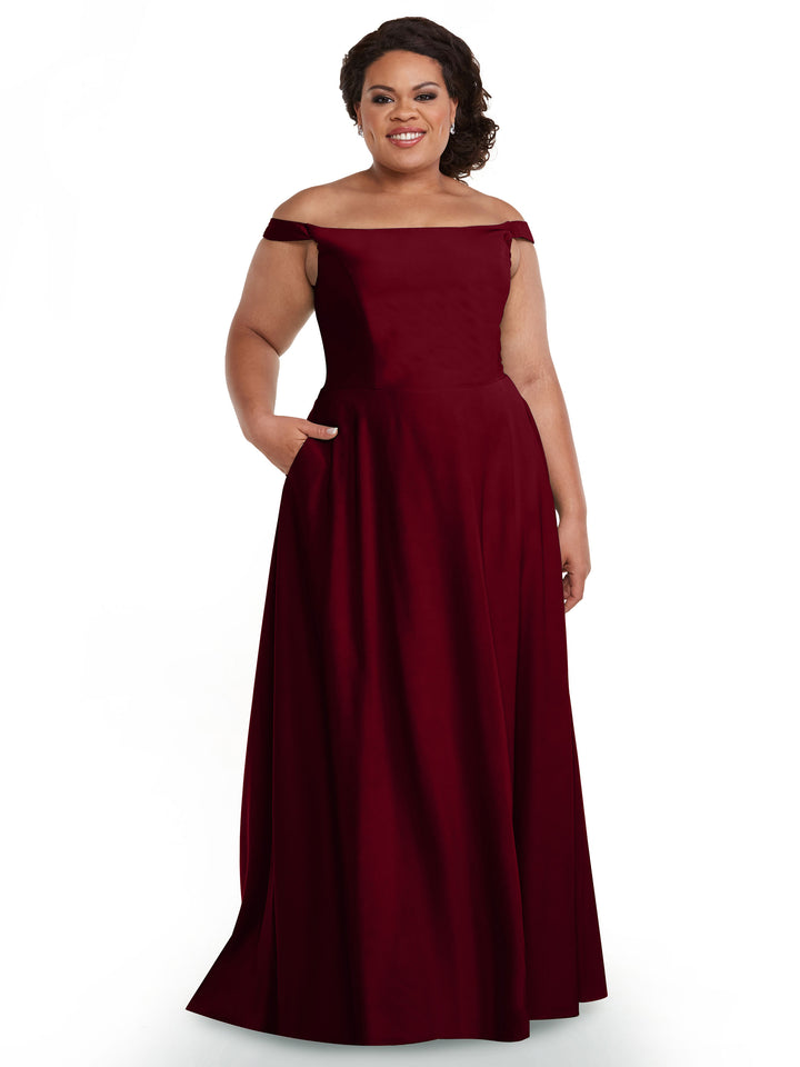 A woman smiles in Avery Austin’s 83005, an elegant deep red off-the-shoulder gown with pockets, a fitted bodice, and flared floor-length skirt. Her dark hair is styled back as she poses against a white background.