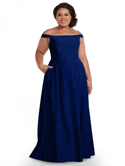 A woman smiles in Avery Austin’s 83005 gown, a dark blue, floor-length dress with an off-the-shoulder neckline, pockets, fitted bodice, and flowing skirt. She poses against a plain white background.