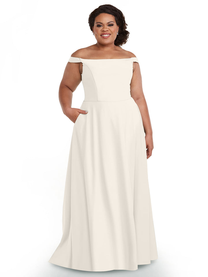 A woman stands smiling in the Avery Austin 83005, a floor-length white off-the-shoulder gown with pockets, against a plain white background.