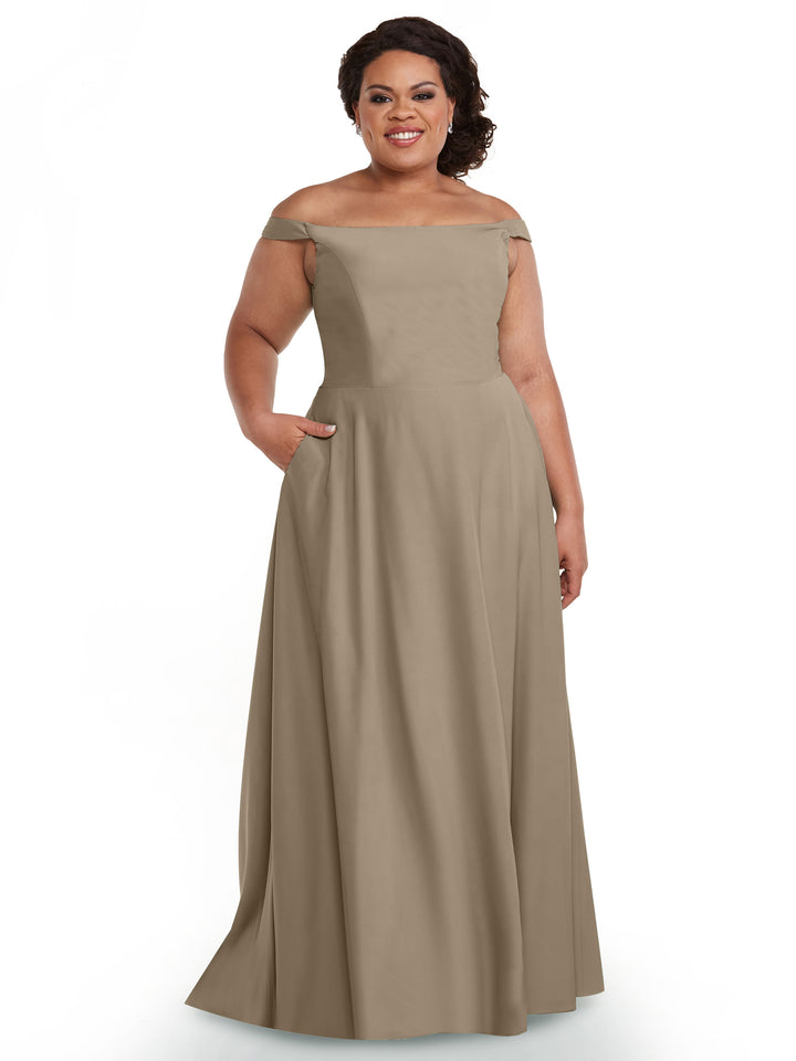 A woman smiles in the Avery Austin 83005 gown, a floor-length taupe off-the-shoulder dress with a straight neckline and pockets, posing against a plain white background.