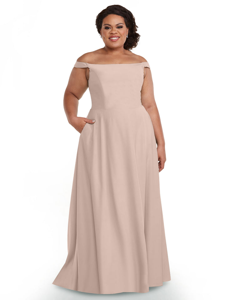 A woman smiles while wearing Avery Austin’s 83005, a rose pink, off-the-shoulder, floor-length dress with pockets, standing against a plain white background.