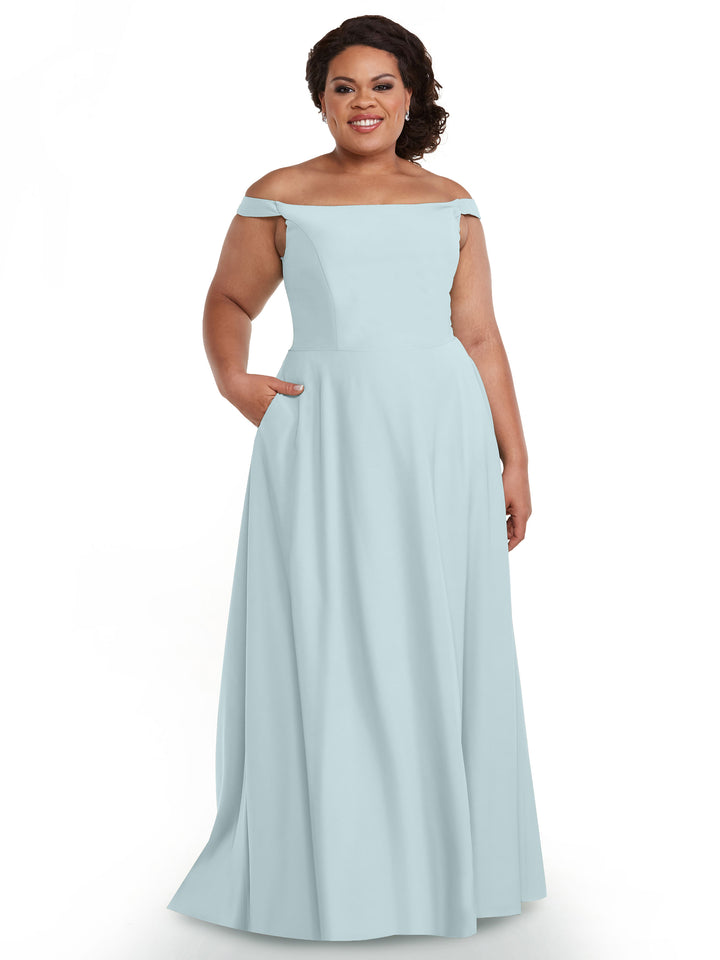 A woman in Avery Austin style 83005, a light blue off-the-shoulder dress with a straight neckline and pockets, stands smiling against a white background.