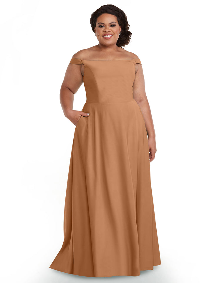A woman smiles in the Avery Austin 83005, a floor-length brown off-the-shoulder dress with a straight neckline and pockets. She poses against a white background, showing off her shoulders and her short, curly dark hair.