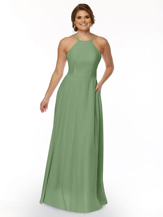 A woman wearing the Avery Austin 83006 sleeveless sage green A-line dress with a halter neckline stands smiling against a white background, her loose waves framing her face and one hand in her pocket.