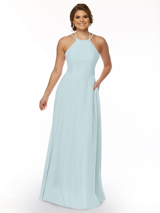 A woman in the Avery Austin 83006, a sleeveless sky blue floor-length A-line dress with a halter neckline, stands against a white background, smiling with one hand in her pocket.