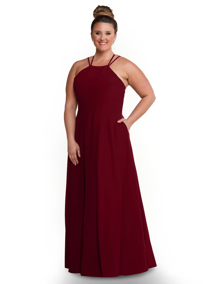 A woman stands and smiles with her hair in a bun, modeling Avery Austin’s 83006—a sleeveless, floor-length burgundy A-line dress with a halter neckline—against a plain white background.