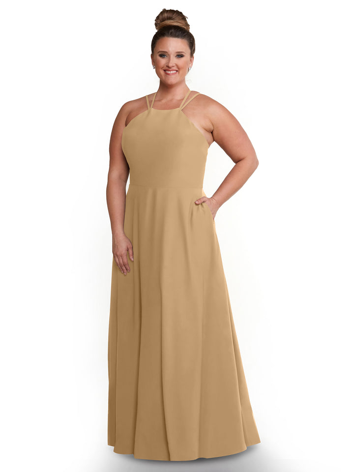 Model wears Avery Austin 83006, a sleeveless, floor-length tan A-line dress with a halter neckline, posing against a white background, smiling with one hand on her hip and her hair in a high bun.