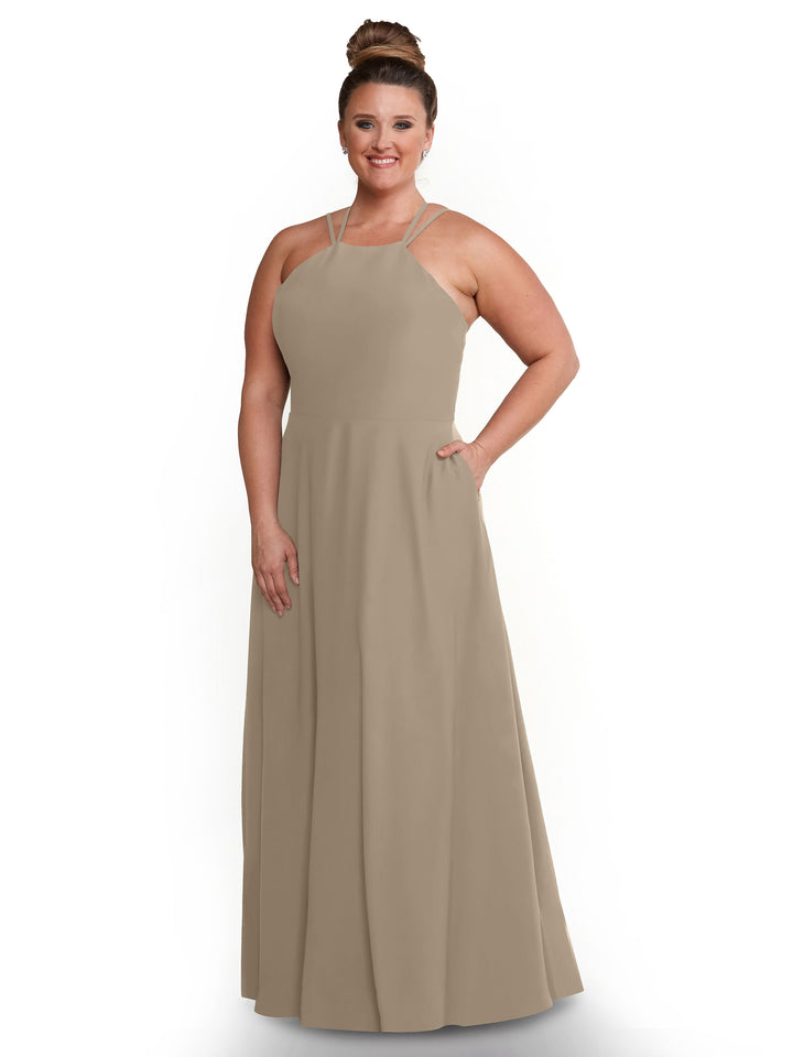 A woman stands smiling against a white background, wearing Avery Austin’s style 83006: a taupe sleeveless floor-length A-Line dress with a halter neckline, her hair in a high bun, one arm by her side and the other resting in a pocket.