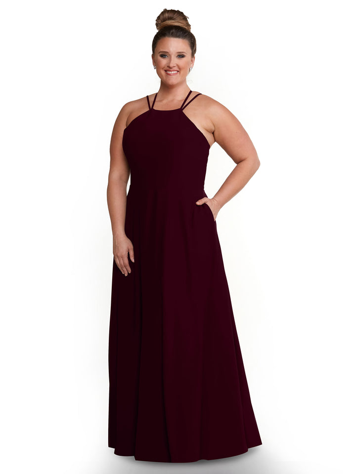 A woman with her hair in a bun models Avery Austin 83006, a sleeveless dark burgundy A-Line dress with double straps and side pockets, against a plain white background.