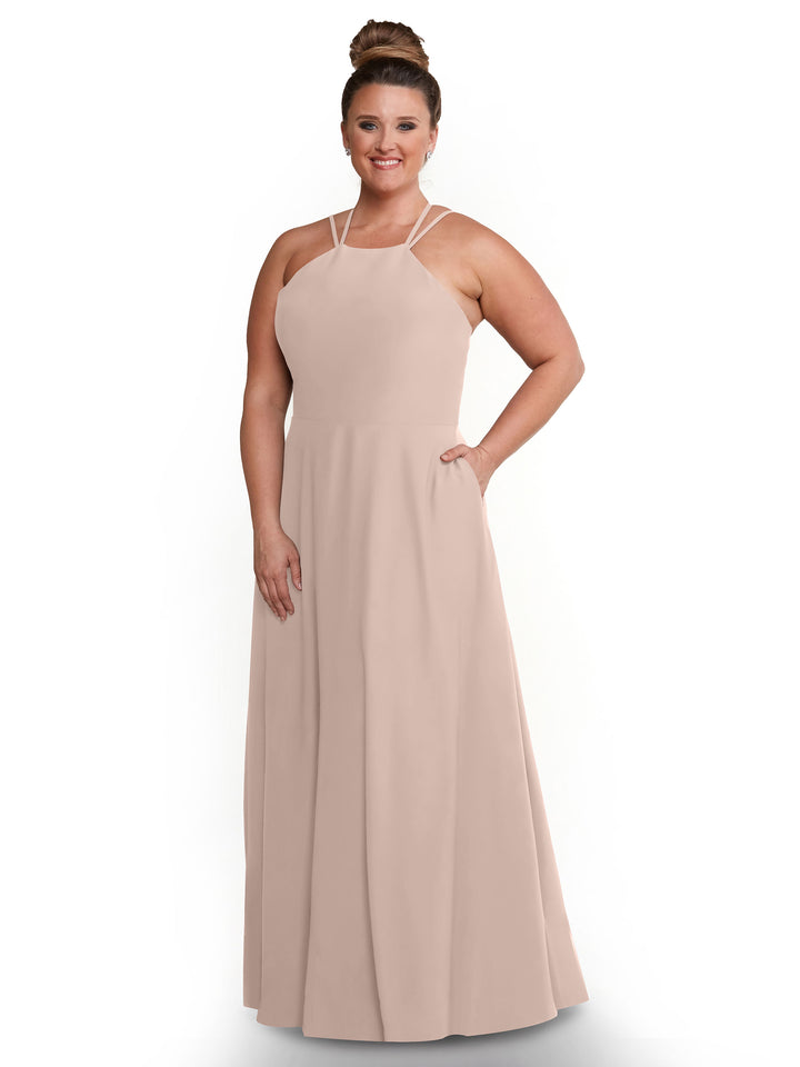 A woman in a rose pink Avery Austin 83006 floor-length A-line dress with a sleeveless halter neckline stands smiling with her hair in a high bun, one hand in her pocket, against a white background.