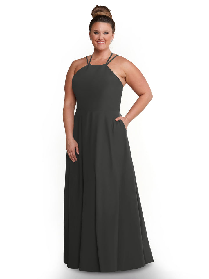 A woman stands smiling with one hand in her pocket, wearing the Avery Austin 83006— a dark gray, floor-length A-line dress with thin straps—against a plain white background.