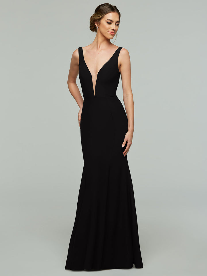 A woman poses against a gray backdrop, wearing Avery Austin’s 83010 sleeveless black mermaid dress with a plunging neckline, her hair in an elegant updo and one hand on her hip.