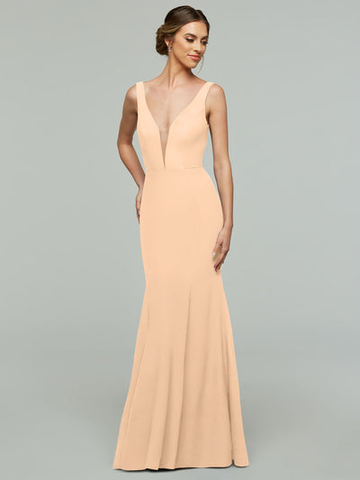 A woman wears Avery Austin’s 83010, a blush mermaid gown with a plunging neckline, sleeveless design, and fitted waist, standing against a plain light gray background.