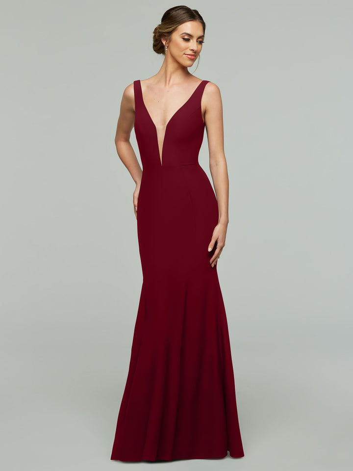 A woman in the Avery Austin 83010, a floor-length burgundy gown with a plunging V-neckline, stands against a plain gray background, gazing to the side with a gentle smile.