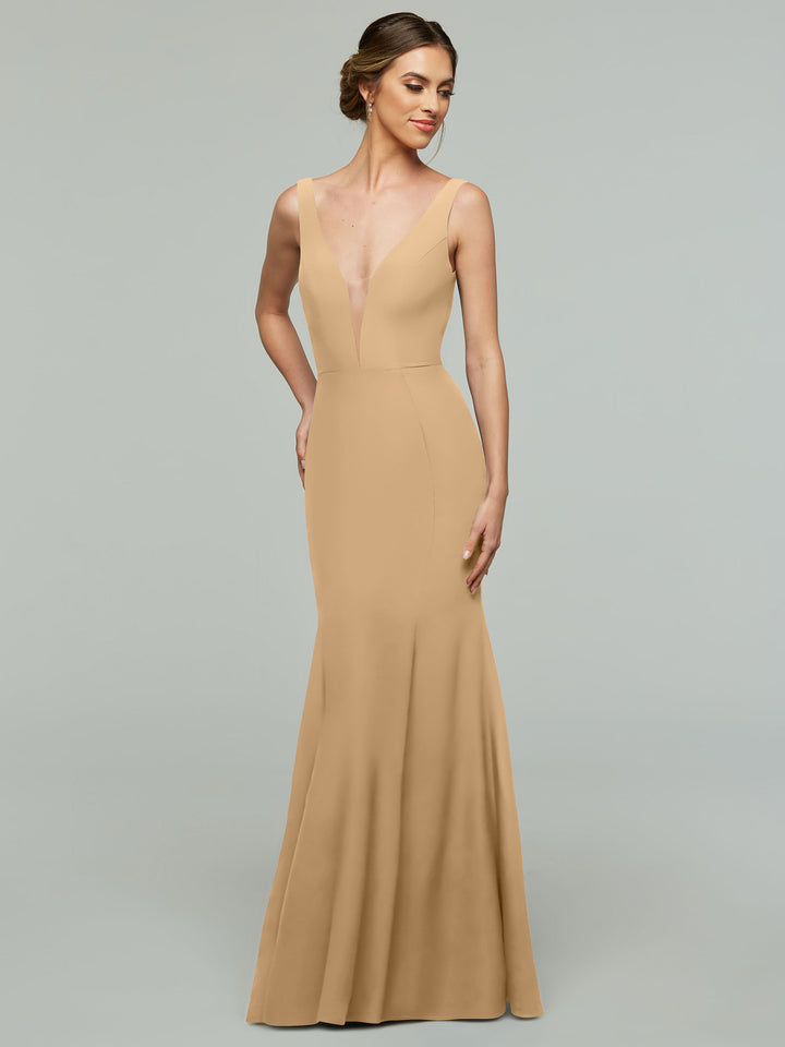 A woman wearing Avery Austin’s 83010 gown in cappuccino, an elegant sleeveless floor-length dress with a plunging neckline, stands before a plain light gray background. Her hair is in an updo and she smiles softly, gazing to the side.